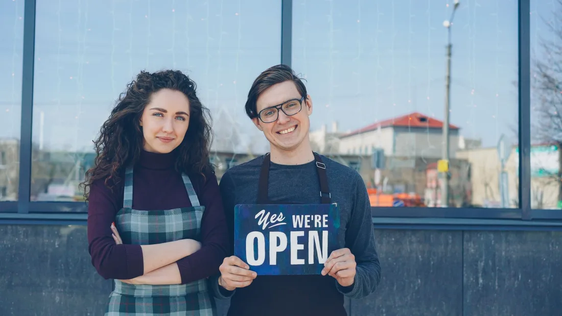 Two cafe owners proudly holding an open sign outside their storefront. Photo by Vitaly Gariev on Pexels