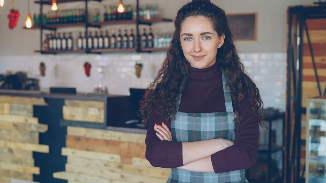 Confident café owner stands with arms crossed inside a rustic, cozy coffee shop. Photo by Vitaly Gariev on Pexels