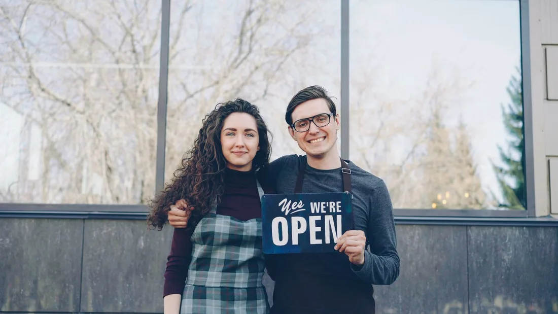 Smiling business owners standing outside with an open sign, ready to welcome customers. Photo by Vitaly Gariev on Pexels