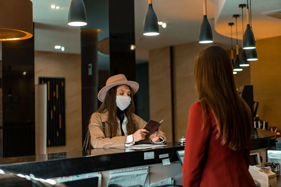 A woman wearing a mask checks in at a hotel reception desk. Photo by Mikhail Nilov on Pexels