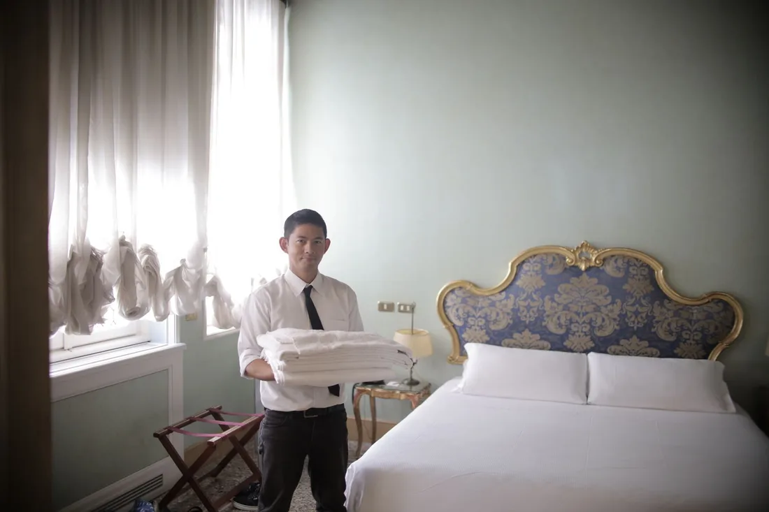 Hotel staff member carrying fresh linens in a bright, neatly made guest room. Photo by Andrea Piacquadio on Pexels