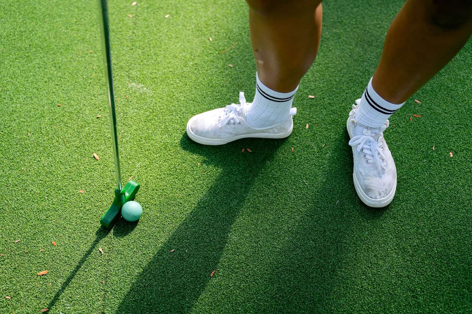 A person in white sneakers standing with a putter and golf ball on a green putting surface. Photo by Anna Tarazevich on Pexels