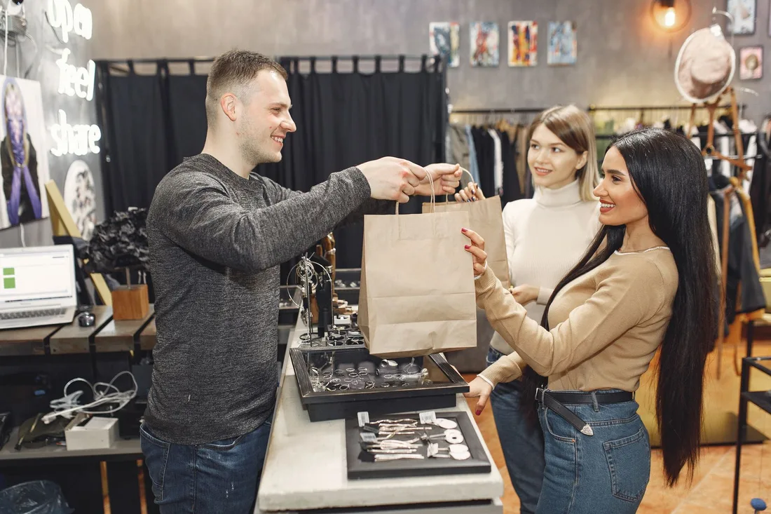 Side view of cheerful male shop assistant standing at counter desk and giving to positive female customers paper bags with purchases in trendy shop. Photo by Gustavo Fring on Pexels