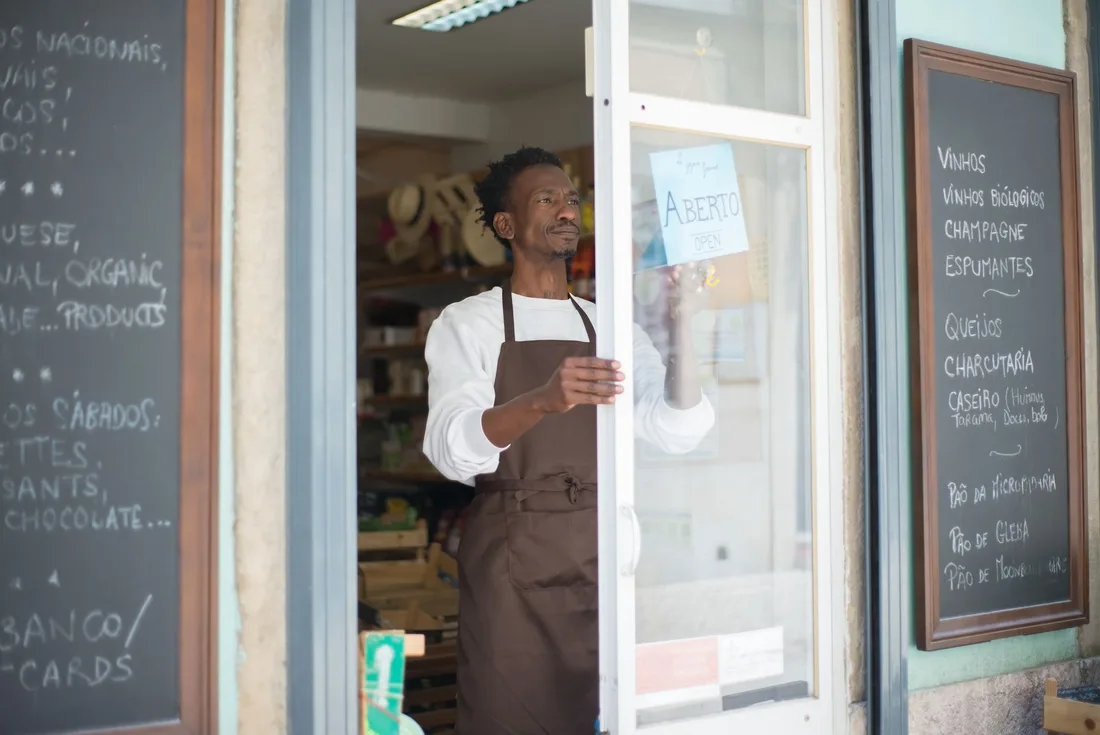 A man in an apron opens a grocery store door in Portugal, ready for business. Photo by Kampus Production on Pexels