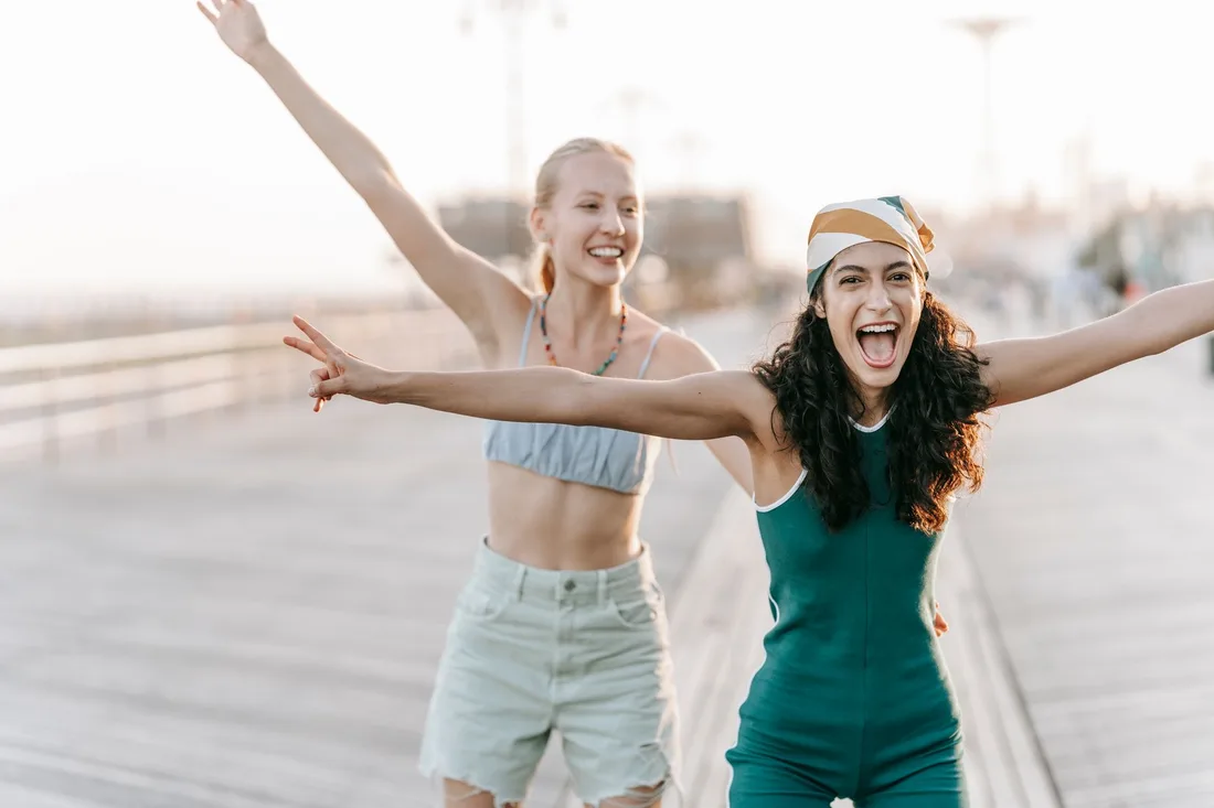 Two joyful women with arms outstretched laughing on a seaside boardwalk. Photo by Katya Wolf on Pexels