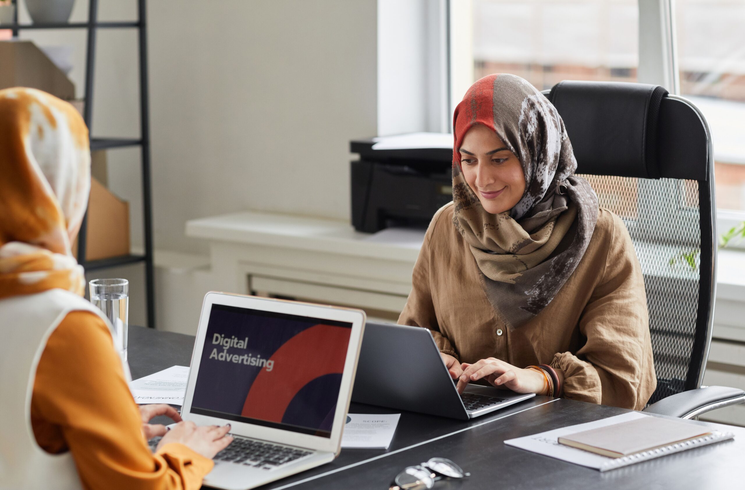Two women in hijabs work on laptops in a bright office. Perfect for business and diversity themes.